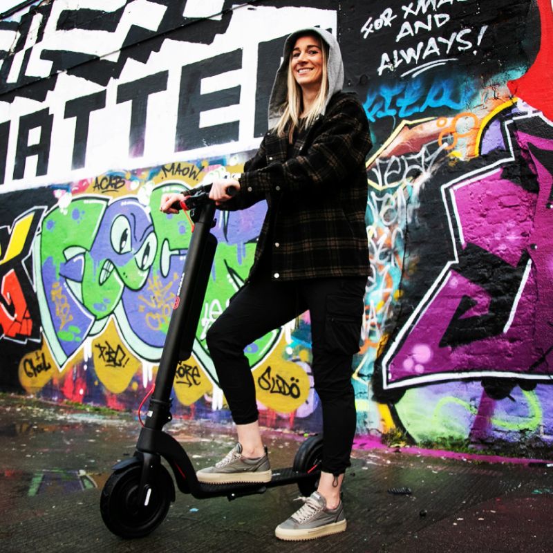 Person riding a Cruzaa Comuta carbon black E-Scooter in front of a colorful graffiti wall.